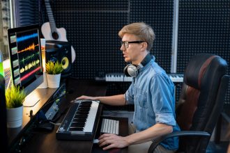 Młody mężczyzna deejay w dżinsowej koszuli patrząc na miksowanie dźwięku na ekranie komputera, siedząc przy biurku przed monitorem