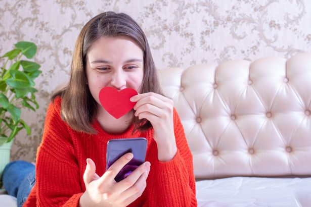 Szczęśliwa młoda dziewczyna w czerwonym swetrze trzyma czerwone papierowe serce i telefon, gratulując rodzinie lub znajomym na Walentynki w domu.