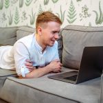 Mężczyzna korzysta z laptopa siedząc na kanapie i pracując zdalnie z domu. Edukacja online na odległość
