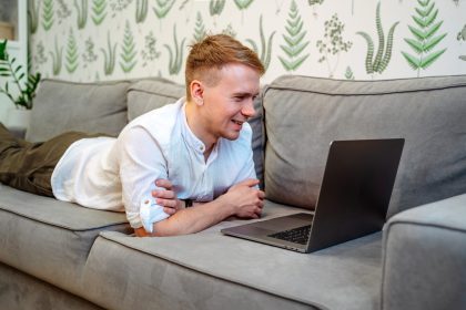Mężczyzna korzysta z laptopa siedząc na kanapie i pracując zdalnie z domu. Edukacja online na odległość
