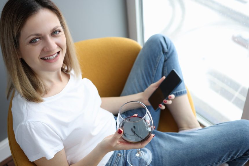 Młoda kobieta siedzi na fotelu przy lampce czerwonego wina i telefonu