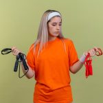 Pewna młoda sportowa dziewczyna rasy kaukaskiej z szelkami w opasce i opaskach trzyma i patrzy na skakanki