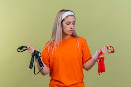 Pewna młoda sportowa dziewczyna rasy kaukaskiej z szelkami w opasce i opaskach trzyma i patrzy na skakanki