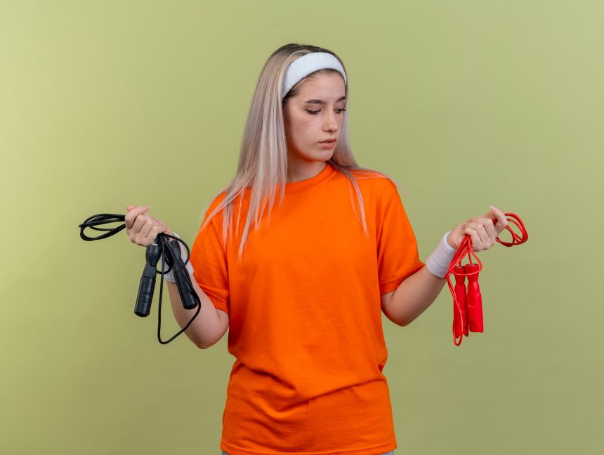 Pewna młoda sportowa dziewczyna rasy kaukaskiej z szelkami w opasce i opaskach trzyma i patrzy na skakanki