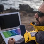 brodaty mężczyzna brodaty podróżnik cyfrowy nomad pracuje z telefonem komórkowym i laptopem na zewnątrz przed oceanem