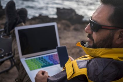 brodaty mężczyzna brodaty podróżnik cyfrowy nomad pracuje z telefonem komórkowym i laptopem na zewnątrz przed oceanem