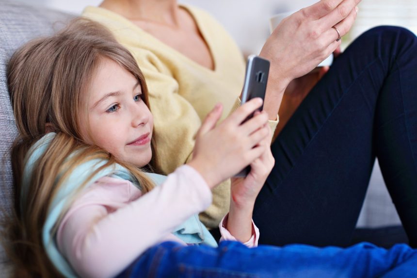 młoda mama i jej córeczka używają smartfona i siedzą na kanapie w domu