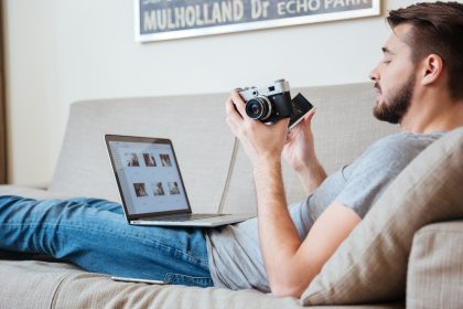 Skoncentrowany, atrakcyjny mężczyzna fotograf używający starego zabytkowego frontu i laptopa na kanapie w domu