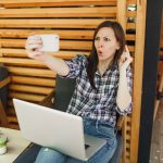 Kobieta w drewnianej kawiarni na zewnątrz ulicy lato siedzi z laptopa komputer pc, robi selfie strzał na telefon komórkowy, relaks w czasie wolnym. Biuro mobilne