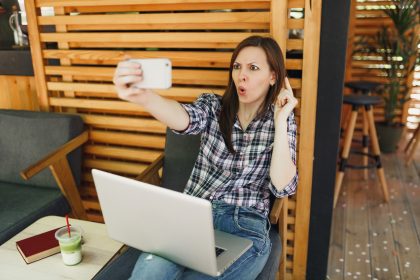 Kobieta w drewnianej kawiarni na zewnątrz ulicy lato siedzi z laptopa komputer pc, robi selfie strzał na telefon komórkowy, relaks w czasie wolnym. Biuro mobilne