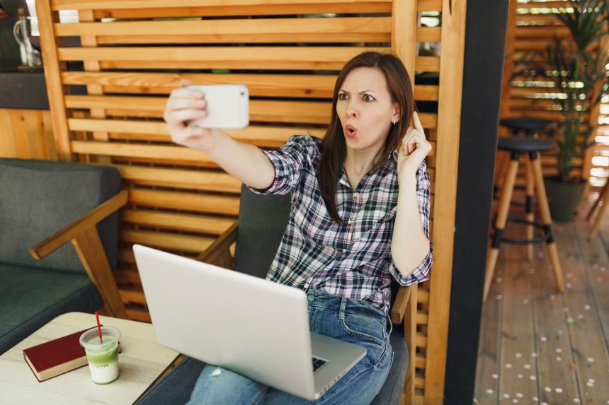 Kobieta w drewnianej kawiarni na zewnątrz ulicy lato siedzi z laptopa komputer pc, robi selfie strzał na telefon komórkowy, relaks w czasie wolnym. Biuro mobilne