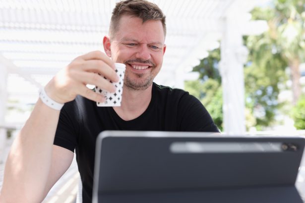Uśmiechnięty młody człowiek siedzący z filiżanką kawy i pracujący na tablecie na ulicy