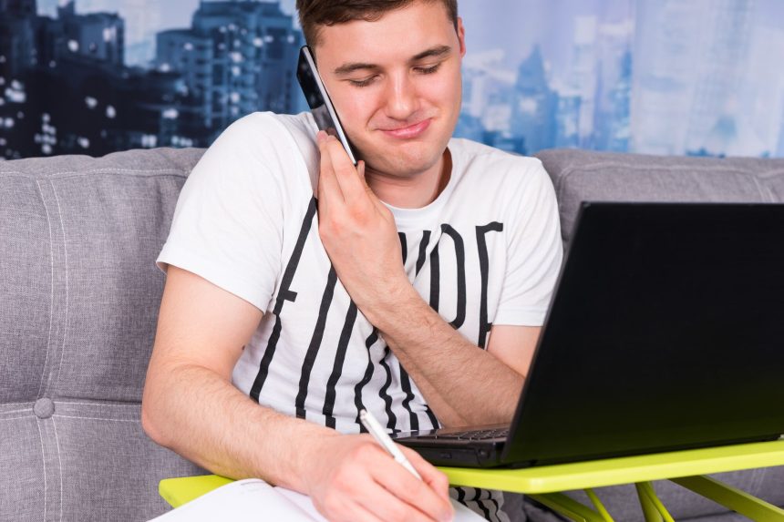 Młody zmęczony mężczyzna pracuje na laptopie, robiąc interesy z domu, siedząc na kanapie, rozmawiając przez telefon komórkowy, trzymając długopis, gdy czyta informacje na ekranie