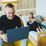 Młody mężczyzna pracuje na laptopie na tle swojej żony, która karmi swoje nowo narodzone dziecko Piersią Praca online na laptopie