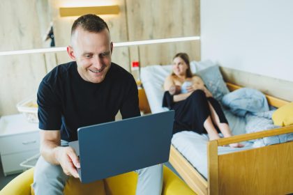 Młody mężczyzna pracuje na laptopie na tle swojej żony, która karmi swoje nowo narodzone dziecko Piersią Praca online na laptopie