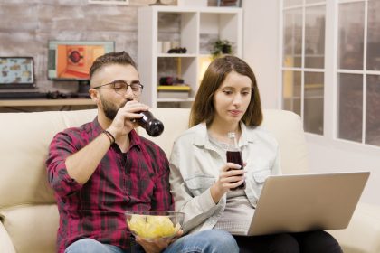 Szczęśliwa młoda para robi zakupy online na laptopie. Para siedzi na kanapie do picia sody i jedzenie frytek.