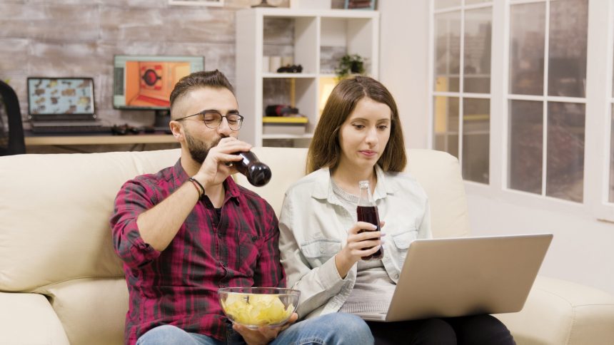 Szczęśliwa młoda para robi zakupy online na laptopie. Para siedzi na kanapie do picia sody i jedzenie frytek.