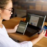 Kobieta rasy mieszanej w okularach pracująca z wieloma elektronicznymi urządzeniami internetowymi Freelancer bizneswoman ma tablet i telefon komórkowy w rękach i laptop na stole z wykresami na ekranie Temat multitasking