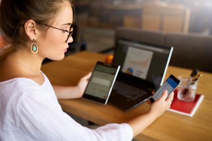 Kobieta rasy mieszanej w okularach pracująca z wieloma elektronicznymi urządzeniami internetowymi Freelancer bizneswoman ma tablet i telefon komórkowy w rękach i laptop na stole z wykresami na ekranie Temat multitasking