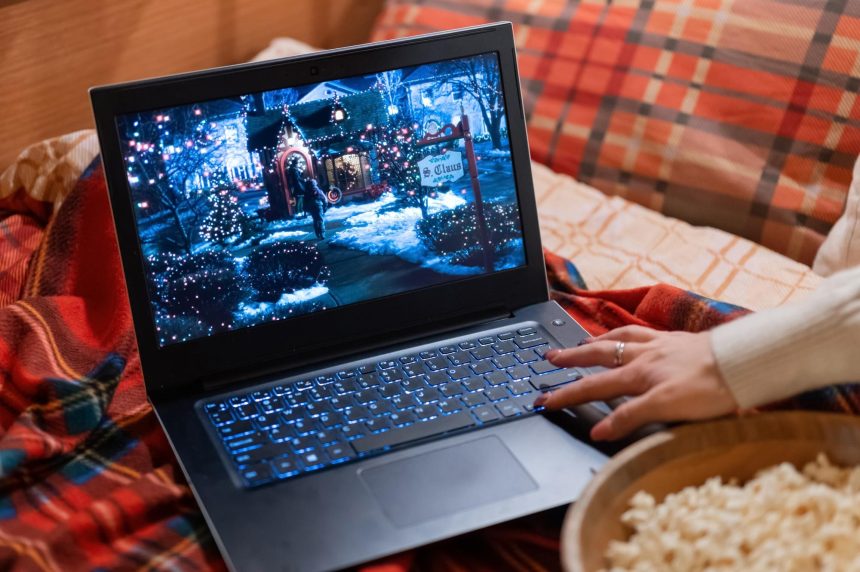 Wyświetlacz laptopa z świątecznym filmem świątecznym „sam w domu” na czerwonej kraciastej kratce w łóżku w domu i kobiecym ręcznym przewijaniem wideo. spędzać czas nadchodząc szczęśliwego nowego roku i wesołych świąt