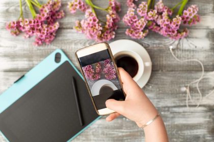 Fotograf Instagram koncepcja warsztatów blogowania, ręka trzyma telefon i robienie zdjęć stylowych kwiatów, filiżankę kawy i tablet graficzny na szarym drewnianym tle rustykalnym. Przestrzeń dla tekstu.