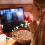 Młoda kobieta ogląda świąteczny film na laptopie z popcornem w domu przyczepa kempingowa w wygodnym łóżku z girlandą w kratę i świecącymi światłami w nocy w wigilię Bożego Narodzenia lub Nowego Roku. ręka na twarzy
