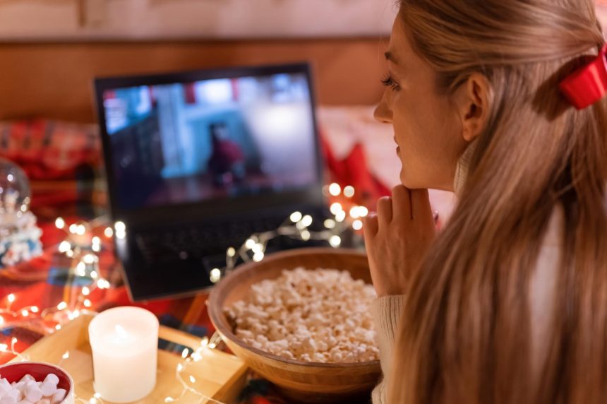 Młoda kobieta ogląda świąteczny film na laptopie z popcornem w domu przyczepa kempingowa w wygodnym łóżku z girlandą w kratę i świecącymi światłami w nocy w wigilię Bożego Narodzenia lub Nowego Roku. ręka na twarzy