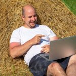 Mężczyzna w średnim wieku, ubrany w T-shirt i szorty, leży na żółtym stogu siana z laptopem w rękach. Praca zdalna, szkolenia. Obszar wiejski. Uśmiech na twojej twarzy