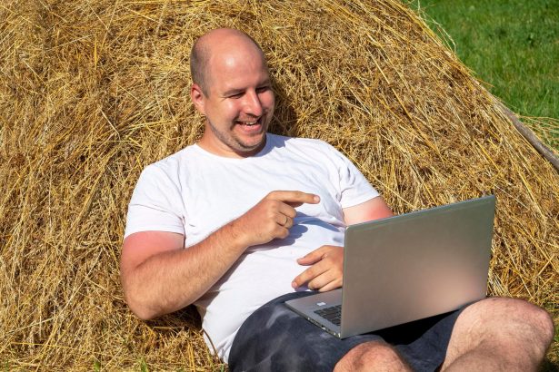 Mężczyzna w średnim wieku, ubrany w T-shirt i szorty, leży na żółtym stogu siana z laptopem w rękach. Praca zdalna, szkolenia. Obszar wiejski. Uśmiech na twojej twarzy