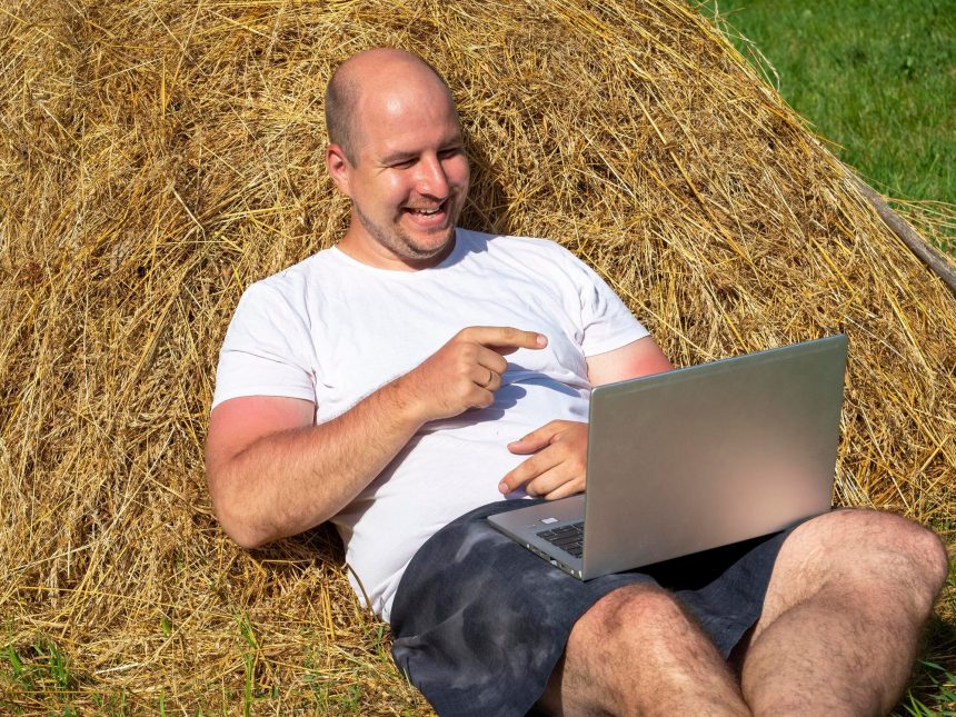 Mężczyzna w średnim wieku, ubrany w T-shirt i szorty, leży na żółtym stogu siana z laptopem w rękach. Praca zdalna, szkolenia. Obszar wiejski. Uśmiech na twojej twarzy