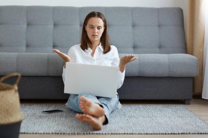 Kryty strzał zdziwionej bezradnej młodej kobiety w białej koszuli i dżinsach, siedzącej na podłodze w pobliżu kaszlu i pracującej na laptopie, patrzącej na wyświetlacz z rozłożonymi ramionami i zdezorientowanym wyrazem twarzy