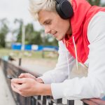 Widok z boku atrakcyjnego mężczyzny rasy kaukaskiej wiadomości za pośrednictwem sieci społecznościowych w smartfonie i za pomocą słuchawek do słuchania muzyki przed jeziorem w parku na świeżym powietrzu Koncepcja stylu życia ludzi