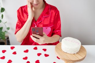 Dziewczyna w czerwonej koszuli używa smartfona, podczas gdy ciasto siedzi na drewnianym stojaku otoczonym w kształcie serca konfetti na stole