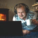 Mężczyzna ogląda w nocy ciekawe treści online na laptopie za pomocą słuchawek w salonie przy kominku