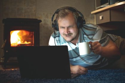 Mężczyzna ogląda w nocy ciekawe treści online na laptopie za pomocą słuchawek w salonie przy kominku