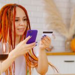 Młoda kobieta z jasnymi dredami z kartą kredytową telefonu komórkowego siedząc w kuchni