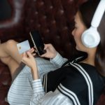 Długowłosa Azjatka siedząca na kanapie w pokoju używa karty kredytowej do dokonywania zakupów za pośrednictwem aplikacji na swoim telefonie komórkowym nosząca białe słuchawki do słuchania muzyki