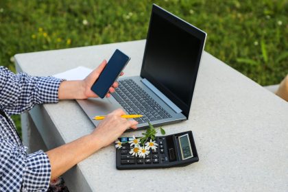 Kobieta pracuje na laptopie i trzyma smartfon w ręku z kalkulatorem leżącym w pobliżu