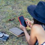 Widok z góry kobiety w czarnej sukience i kapeluszu patrzącej na smartfon leżący na brzuchu