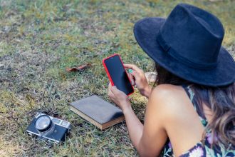 Widok z góry kobiety w czarnej sukience i kapeluszu patrzącej na smartfon leżący na brzuchu