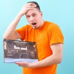 zszokowany mężczyzna trzymający głowę i otwarty brudny i zakurzony laptop na niebiesko. Przegrzanie
