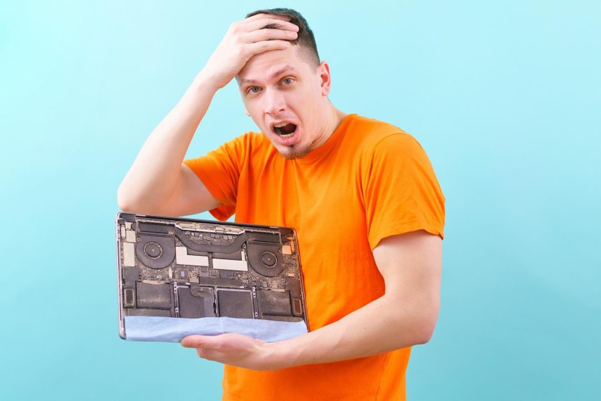 zszokowany mężczyzna trzymający głowę i otwarty brudny i zakurzony laptop na niebiesko. Przegrzanie