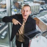 Portret młodego radosnego mężczyzny stojącego w pobliżu okna z laptopem w rękach Praca w biurze Udany menedżer płci męskiej