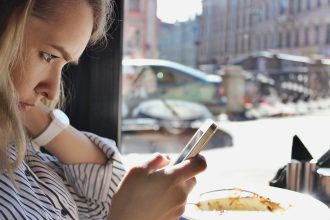 Profil młodej jasnowłosej kobiety korzystającej z telefonu komórkowego lub gadżetu mobilnego siedzącego w kawiarni