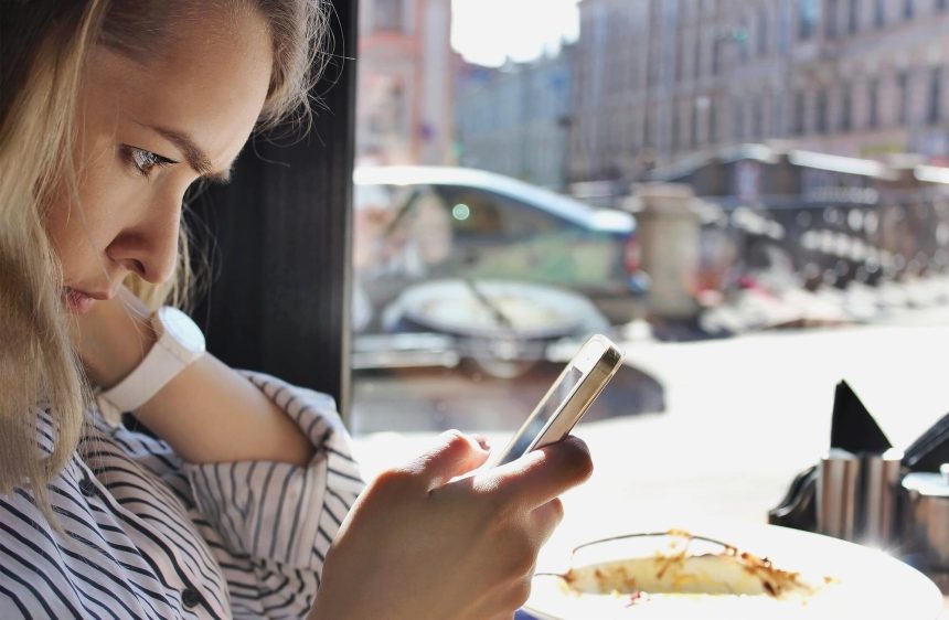 Profil młodej jasnowłosej kobiety korzystającej z telefonu komórkowego lub gadżetu mobilnego siedzącego w kawiarni