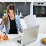 Nastolatka trzymająca papiery i robiąca notatki na nalepce przed laptopem w salonie w domu