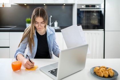 Nastolatka trzymająca papiery i robiąca notatki na nalepce przed laptopem w salonie w domu