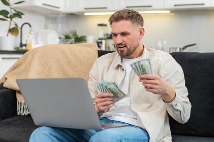 Młody mężczyzna liczy gotówkę w dolarach, porównuje z arkuszem zarobków na laptopie, wpisuje sumy i uśmiecha się z powodu osiągnięcia celu.