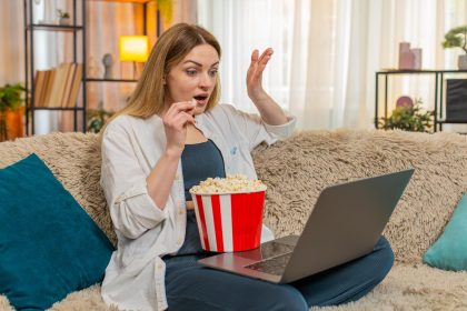 Uśmiechnięta kobieta ogląda film na laptopie w domu, je popcorn i reaguje emocjonalnie, okazując radość