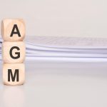Three wood cubes stacked with letters agm placed in front of a stack of documents on a desk clean